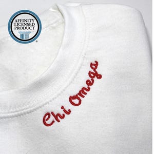 May include: White sweatshirt with the embroidered text "Chi Omega" in red script. The sweatshirt has a crew neck and a round, blue and white "Affinity Licensed Product" logo.