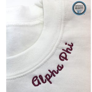Op de afbeelding: Close-up van een witte sweatshirt met de geborduurde woorden "Alpha Phi" in een bordeauxrood script. De sweatshirt heeft een ronde hals en een label met "Affinity Licensed Product".