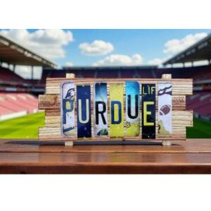 May include: A wooden sign spelling "PURDUE" with license plates. The sign is set against a backdrop of a stadium with a green field and blue sky. The letters are in various colors.