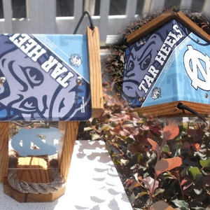 May include: Wooden birdhouse featuring the University of North Carolina Tar Heels logo and colors. The birdhouse has a light blue roof with the words "TAR HEELS" and the UNC logo. The front has a clear panel with the UNC logo and a rope perch.