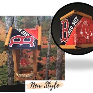 May include: A wooden bird feeder with a red, white, and blue Boston Red Sox logo. The feeder has a rope perch and a clear plastic bottom. The feeder is hanging from a tree branch.
