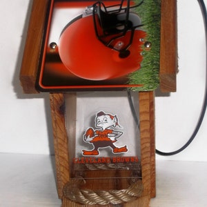 Może przedstawiać: Drewniany karmnik dla ptak&oacute;w z logo Cleveland Browns i małą figurką elfa trzymającego piłkę nożną. Karmnik ma przezroczyste plastikowe okno i sznurkowy percz.