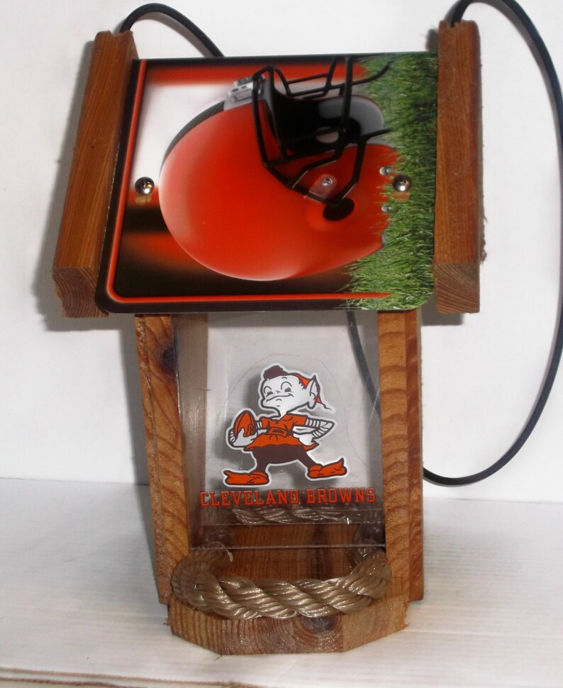 Może przedstawiać: Drewniany karmnik dla ptak&oacute;w z logo Cleveland Browns i małą figurką elfa trzymającego piłkę nożną. Karmnik ma przezroczyste plastikowe okno i sznurkowy percz.