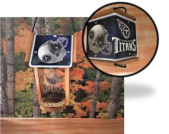May include: A wooden bird feeder with a Tennessee Titans logo. The feeder has a clear plastic container and a metal roof. The roof is decorated with a Tennessee Titans logo and a football helmet.