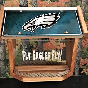 Może przedstawiać: Drewniane karmnik dla ptaków z przezroczystym plastikowym przodem i zielono-białym logo Philadelphia Eagles na górze. Karmnik ma sznurkowy drążek i napis "FLY EAGLES FLY!" nadrukowany z przodu.