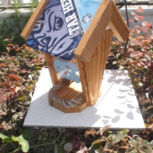 May include: Wooden birdhouse with a pitched roof made from license plates. The plates feature the words "TAR HEEL" and a logo. The birdhouse has a light-colored wood frame and a decorative rope detail.