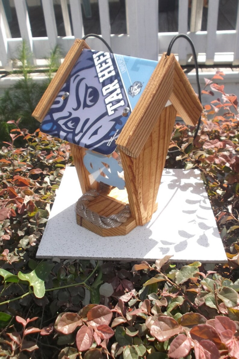 May include: Wooden birdhouse with a pitched roof made from license plates. The plates feature the words "TAR HEEL" and a logo. The birdhouse has a light-colored wood frame and a decorative rope detail.