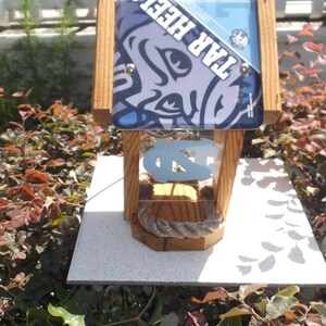 May include: Wooden birdhouse with a Tar Heels theme. The birdhouse features a light blue and navy blue design with the words "TAR HEELS" and the UNC logo. The birdhouse is made of wood and has a rope detail.