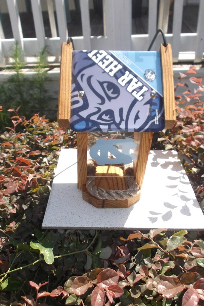 May include: Wooden birdhouse with a Tar Heels theme. The birdhouse features a light blue and navy blue design with the words "TAR HEELS" and the UNC logo. The birdhouse is made of wood and has a rope detail.