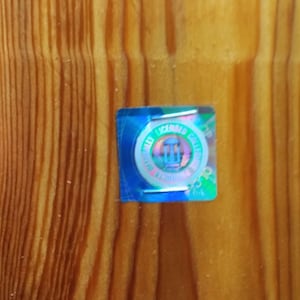 May include: A small, iridescent sticker with the text "MAX LICENSED COLLECTIBLES" and a central emblem, affixed to a wooden surface with vertical grain. The sticker has a blue border and a holographic effect.