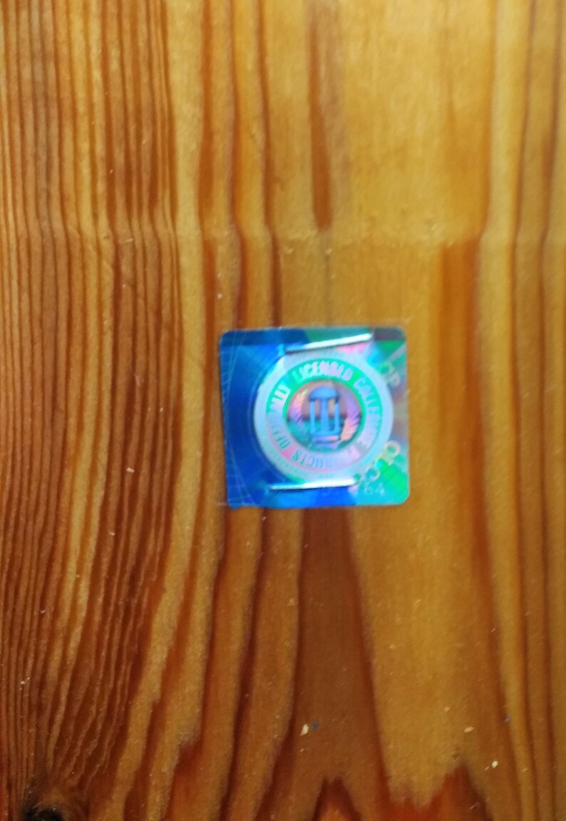 May include: A small, iridescent sticker with the text "MAX LICENSED COLLECTIBLES" and a central emblem, affixed to a wooden surface with vertical grain. The sticker has a blue border and a holographic effect.