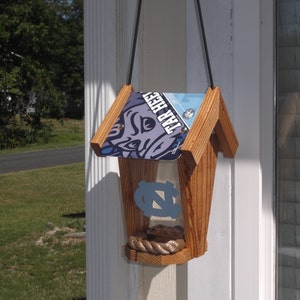 May include: Wooden birdhouse with a license plate roof featuring the text "TAR HEEL" and a Carolina blue logo. The birdhouse has a rope perch and a clear front panel. A black cord is used for hanging.
