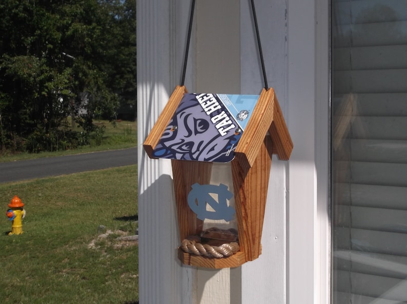 May include: Wooden birdhouse with a license plate roof featuring the text "TAR HEEL" and a Carolina blue logo. The birdhouse has a rope perch and a clear front panel. A black cord is used for hanging.