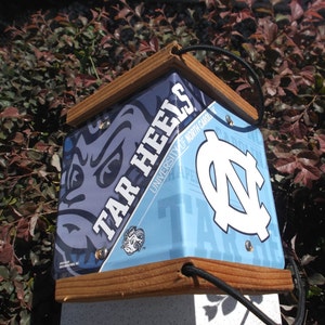 May include: A square birdhouse with a wooden frame and a blue and white design featuring the words "TAR HEELS" and the University of North Carolina logo. The birdhouse has a black cord for hanging.