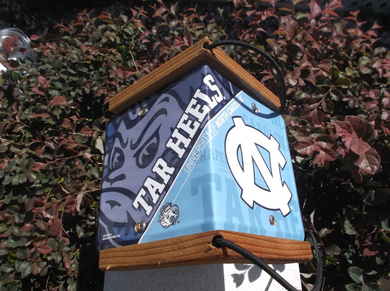 May include: A square birdhouse with a wooden frame and a blue and white design featuring the words "TAR HEELS" and the University of North Carolina logo. The birdhouse has a black cord for hanging.