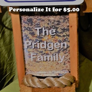 May include: A wooden bird feeder with a clear front panel filled with birdseed. The text "The Pridgen Family" is displayed on the panel. The feeder has a rope detail at the bottom and is framed with orange and natural wood.