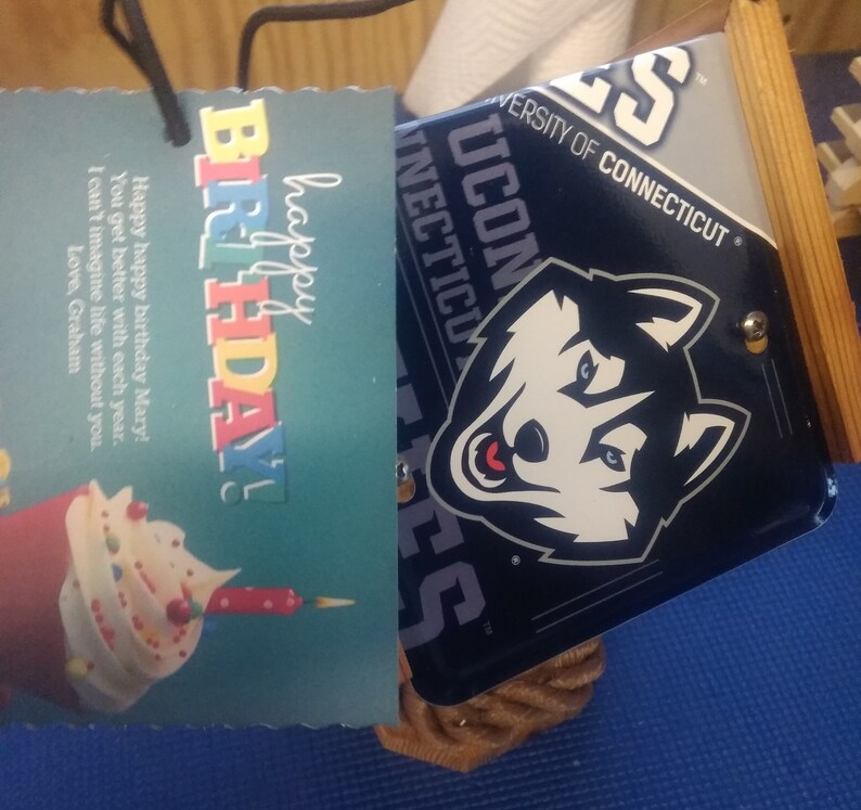 Może przedstawiać: Niebieska i biała kartka urodzinowa z tekstem "happy birthday!" i babeczką z świeczką. Kartka znajduje się obok niebieskiego i białego logo University of Connecticut Huskies z tekstem "University of Connecticut Huskies".