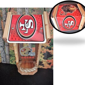 Może przedstawiać: Drewniany karmnik dla ptaków z logo San Francisco 49ers. Karmnik ma czerwono-złoty design z logo drużyny. Zawiera sznurkową grzędę i przezroczysty plastikowy przód.