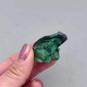 May include: A small, carved frog figurine in shades of green and black, with a touch of purple. The frog is held in a hand against a gray background. The figurine is likely made of stone or a similar material.