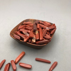 May include: A wooden bowl filled with red jasper tumbled stones. The bowl is sitting on a gray surface. There are several red jasper stones scattered around the bowl.
