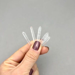May include: A hand holding a cluster of clear quartz crystal points against a gray background. The crystals are transparent and have a pointed shape. The hand has polished nails.