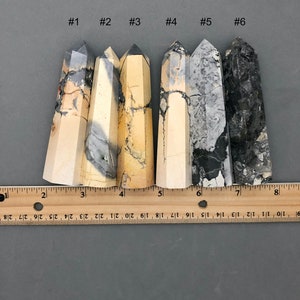 K&ouml;nnte beinhalten: Sechs polierte, spitze, nat&uuml;rliche Stein-Kristallst&auml;be in verschiedenen Graut&ouml;nen, Schwarz und Creme. Die St&auml;be sind von links nach rechts in einer Reihe angeordnet und von 1 bis 6 nummeriert. Ein Lineal befindet sich unter den St&auml;ben als Gr&ouml;&szlig;enreferenz.