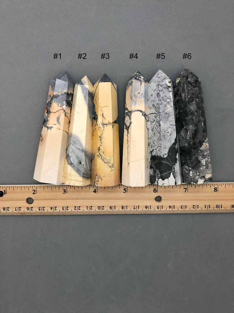 K&ouml;nnte beinhalten: Sechs polierte, spitze, nat&uuml;rliche Stein-Kristallst&auml;be in verschiedenen Graut&ouml;nen, Schwarz und Creme. Die St&auml;be sind von links nach rechts in einer Reihe angeordnet und von 1 bis 6 nummeriert. Ein Lineal befindet sich unter den St&auml;ben als Gr&ouml;&szlig;enreferenz.