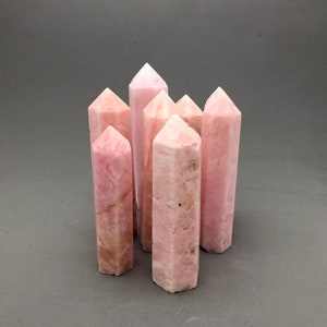 May include: A group of six pink crystal points, also known as pink calcite, arranged in a cluster. The points are all different sizes and shapes, but they are all a similar shade of pink.