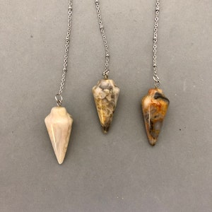 May include: Three gemstone pendulums with silver chains hang against a gray background. The pendulums are cone-shaped, each with a unique pattern of colors including cream, brown, and gold. The chains are delicate and attached at the top of each pendulum.