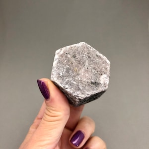 May include: A hexagonal shaped, grey and white, rough-textured, natural gemstone.