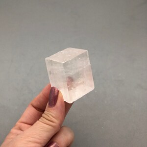 May include: A clear, translucent, rectangular selenite crystal with smooth, polished faces. The crystal is held in a hand.