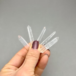 May include: A collection of clear quartz crystal points, each with a pointed tip. The crystals are held against a gray background. The crystals are transparent and appear to be natural formations.