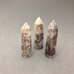 May include: Three white crystal points with purple and brown streaks. The points are arranged in a row on a gray surface.