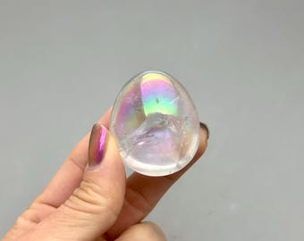 May include: A clear, egg-shaped crystal with iridescent rainbow hues. The crystal has a smooth, polished surface and appears to be held in a hand. The background is a solid gray.