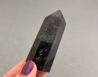 May include: A dark grey crystal point with a pointed tip and a smooth, polished surface. The crystal has a cloudy, translucent appearance with flecks of gold throughout.