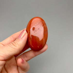 May include: A smooth, oval-shaped red jasper stone with a polished finish. The stone has a deep red color with subtle brown and white markings.