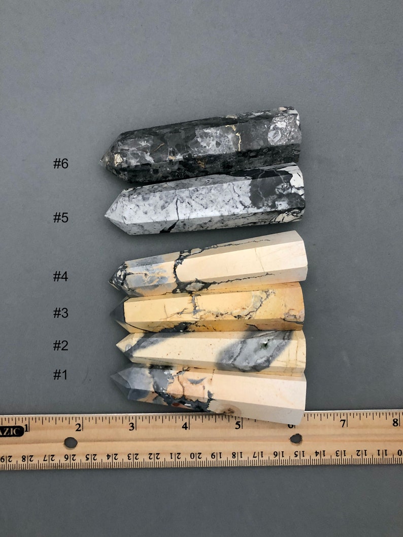 K&ouml;nnte beinhalten: Sechs polierte, spitze, nat&uuml;rliche Stein Zauberst&auml;be, nummeriert von 1 bis 6, vertikal auf einer grauen Oberfl&auml;che angeordnet. Die Zauberst&auml;be sind in verschiedenen Beige-, Wei&szlig;- und Graut&ouml;nen mit schwarzen und braunen Markierungen. Ein Holzlineal befindet sich am unteren Rand des Bildes.