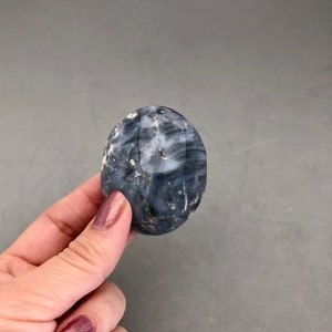 May include: A polished, oval-shaped stone with a mix of dark blue and white marbling. The stone is held in a hand with polished nails, and the background is a solid gray.