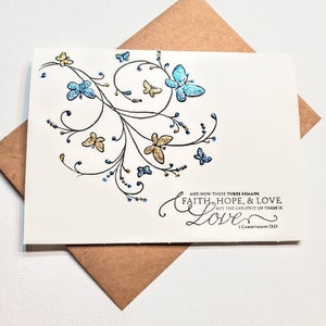 May include: A white card with a black and blue floral design with glitter accents. The card has a quote from the Bible, 1 Corinthians 13:13, "And now these three remain: faith, hope, and love. But the greatest of these is love."