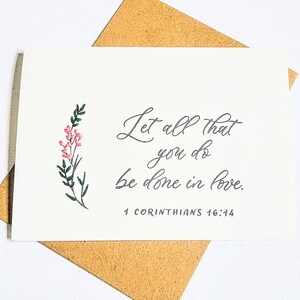 May include: A white greeting card with the handwritten text "Let all that you do be done in love." A floral illustration with pink and green accents is on the left. The card is on a brown envelope. The Bible verse 1 Corinthians 16:14 is at the bottom.