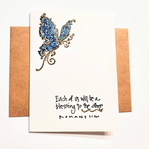 May include: A greeting card with a blue and gold glitter butterfly design. The card includes the text "Each of us will be a blessing to the other" and "Romans 1:12". The card is paired with a brown envelope.