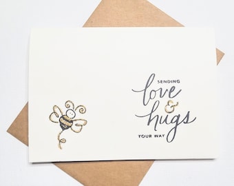 May include: A cream-colored greeting card with a gold glitter bee illustration and the words "Sending love & hugs your way" in black script. The card is inside a brown envelope.