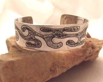 May include: A silver cuff bracelet with a textured, wave-like black design. The bracelet is displayed on a light brown, textured surface. The open cuff design allows for easy wear. This piece is a stylish accessory.