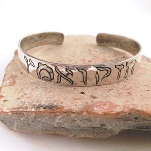 May include: A silver cuff bracelet with etched Hebrew text. The bracelet has a hammered texture and is displayed on a light brown, textured surface. The open-ended design allows for easy wear.