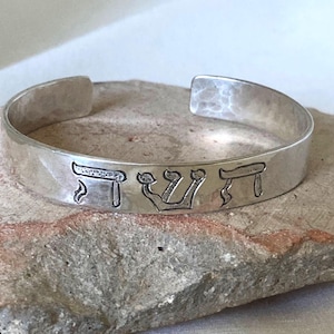 May include: A silver cuff bracelet with a hammered texture and an open back. The front of the band features three engraved Hebrew characters, specifically the letters Shin, Vav, and Shin, which spell out the word Shaddai in a dark, recessed font.