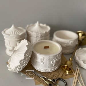 May include: White ceramic candle holders shaped like a carousel with a white candle in one of the holders. The candle holders have intricate details and are surrounded by gold scissors, wooden matches, and a gold metal bell.