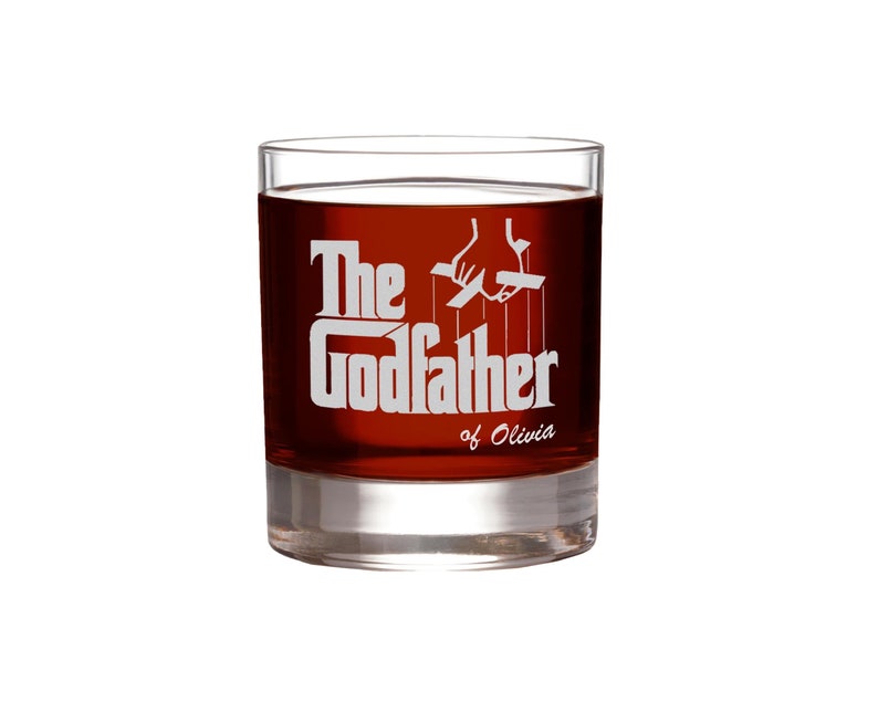 May include: A clear glass tumbler with a red liquid inside. The glass has a white design that says "The Godfather of Olivia" with a silhouette of a hand controlling strings.