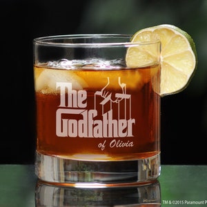 May include: A clear glass with a brown liquid and ice cubes. The glass has the text "The Godfather of Olivia" etched on the side. A lime wedge is on the rim of the glass.