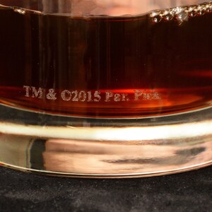 May include: A clear glass tumbler with a brown liquid inside. The bottom of the glass has the text "TM & C2015 Per. Pic."