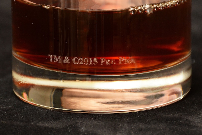 May include: A clear glass tumbler with a brown liquid inside. The bottom of the glass has the text "TM & C2015 Per. Pic."